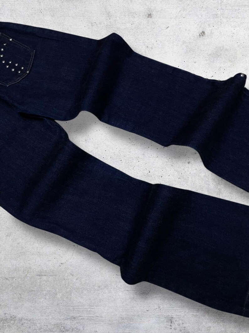 Puede incluir: Vaqueros de mezclilla azul oscuro con un dise&ntilde;o cl&aacute;sico de cinco bolsillos. El bolsillo trasero presenta un patr&oacute;n decorativo de peque&ntilde;os tachuelas plateadas. Los vaqueros est&aacute;n doblados y colocados planos, mostrando la textura y el color de la tela.
