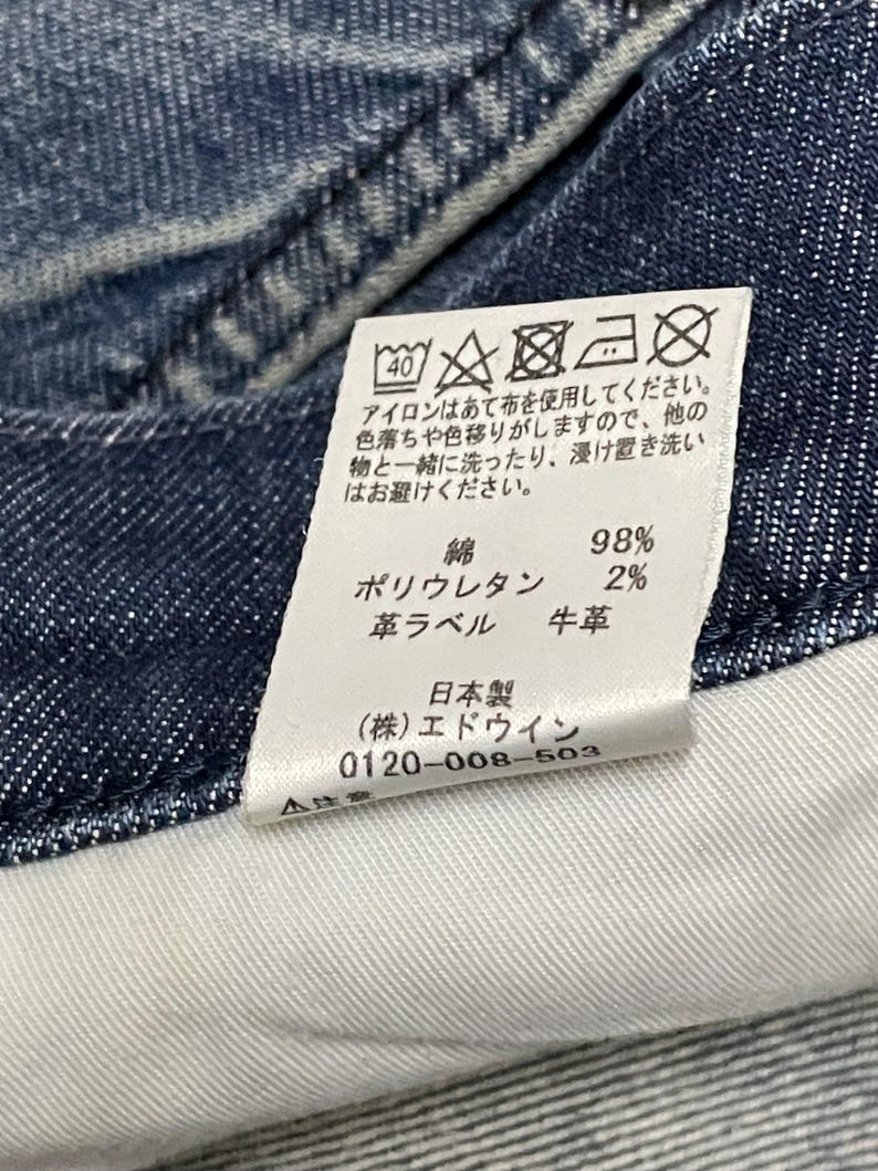 以下が含まれることがあります： デニム生地のクローズアップ。白いタグには洗濯表示と生地組成が表示されています。綿98％、ポリウレタン2％、革ラベル。タグのテキストは日本語です。