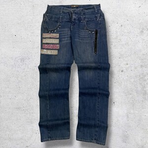 Op de afbeelding: Blauwe spijkerbroek met decoratieve patches en een ritssluiting. De jeans heeft een dubbele knoopsluiting en klinknagels op de zakken. Patches bevatten tekst en gestreepte ontwerpen. De jeans hebben een medium wassing.