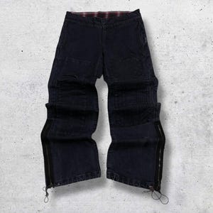 May include: Dark blue denim pants with a plaid lining at the waist. The pants have a unique design with multiple panels and zippers along the outer seams. The bottom of the legs have drawstrings.