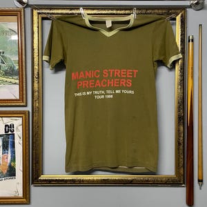 May include: Olive green V-neck t-shirt with red text that reads "MANIC STREET PREACHERS THIS IS MY TRUTH, TELL ME YOURS TOUR 1998". The shirt has a light green trim around the collar and sleeves. The shirt is hanging on a clothesline.
