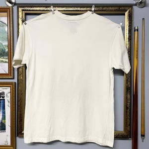May include: A plain cream-coloured, short-sleeved t-shirt hanging on a clothesline. The shirt is made of cotton. The shirt is hanging in front of a framed picture and two pool cues.