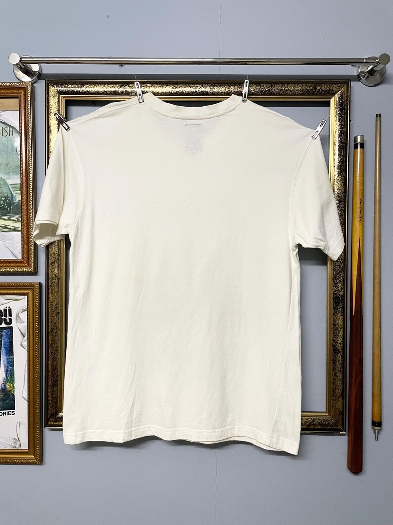 May include: A plain cream-coloured, short-sleeved t-shirt hanging on a clothesline. The shirt is made of cotton. The shirt is hanging in front of a framed picture and two pool cues.