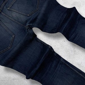 Op de afbeelding: Donkerblauwe denim jeans met zichtbare stiksels en achterzakken. De jeans is opgevouwen, wat de textuur en kleurvariaties van de stof laat zien. De denim lijkt een klassiek, duurzaam materiaal te zijn.