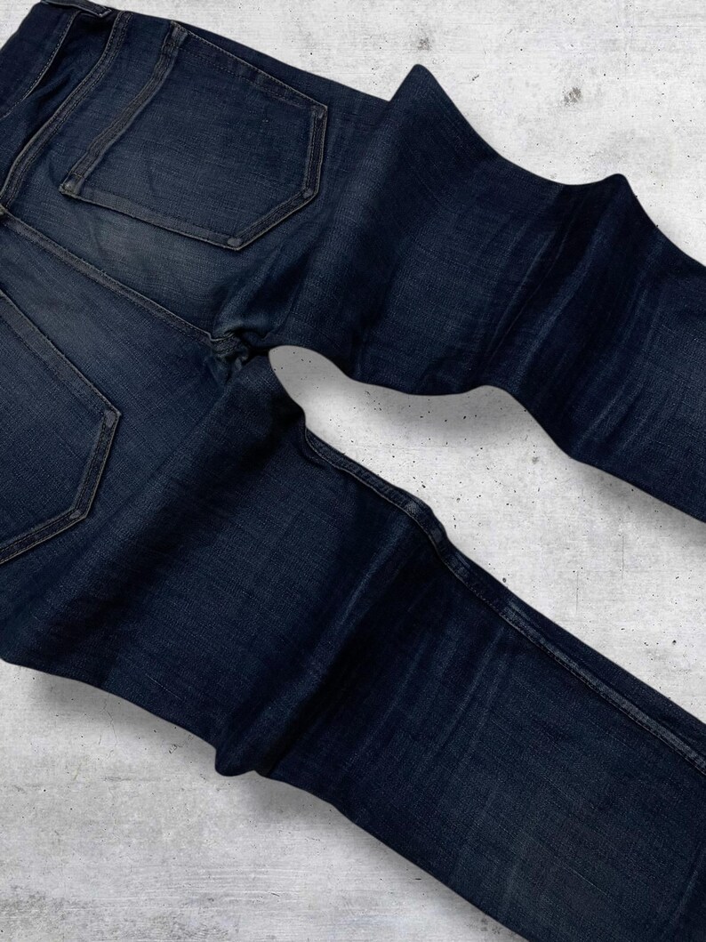 Op de afbeelding: Donkerblauwe denim jeans met zichtbare stiksels en achterzakken. De jeans is opgevouwen, wat de textuur en kleurvariaties van de stof laat zien. De denim lijkt een klassiek, duurzaam materiaal te zijn.