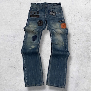 Könnte beinhalten: Verblasste blaue Jeans mit mehreren Aufnähern und Abnutzungserscheinungen. Zu den Merkmalen gehören ein großer blauer Aufnäher am linken Knie, verschiedene kleinere Aufnäher und ein Used-Look. Die Jeans haben ein klassisches Fünf-Taschen-Design.