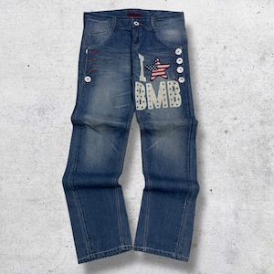 Puede incluir: Vaqueros azules con un lavado descolorido. Los vaqueros presentan detalles de botones blancos y un parche bordado con una estrella y la bandera estadounidense. El texto "I BMB" también está bordado en la parte delantera.