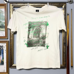 May include: Cream-coloured t-shirt featuring a green and grey graphic print and text. The text includes the phrases "It's up to human beings to protect" and "with good people's hands". The t-shirt is hanging on a metal bar.