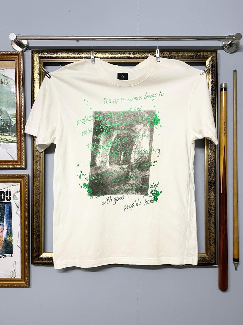 May include: Cream-coloured t-shirt featuring a green and grey graphic print and text. The text includes the phrases "It's up to human beings to protect" and "with good people's hands". The t-shirt is hanging on a metal bar.