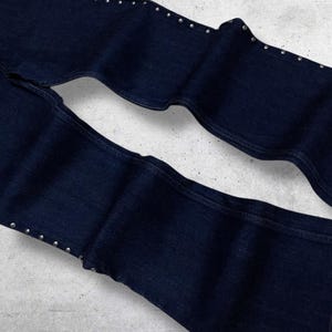Puede incluir: Vaqueros de mezclilla azul oscuro con tachuelas plateadas a lo largo de las costuras exteriores. Los vaqueros est&aacute;n doblados, mostrando los detalles de la tela y la colocaci&oacute;n de las tachuelas. La mezclilla es de un color &iacute;ndigo profundo cl&aacute;sico.