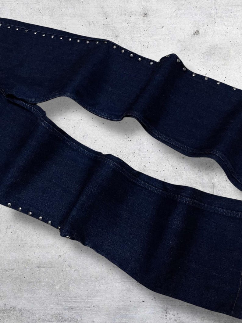 Puede incluir: Vaqueros de mezclilla azul oscuro con tachuelas plateadas a lo largo de las costuras exteriores. Los vaqueros est&aacute;n doblados, mostrando los detalles de la tela y la colocaci&oacute;n de las tachuelas. La mezclilla es de un color &iacute;ndigo profundo cl&aacute;sico.