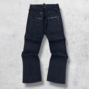 以下が含まれることがあります： ユニークなデザイン要素を備えた濃いネイビーブルーのデニムパンツ。ジッパーポケット、金属製のハードウェア、補強された膝パネルが特徴です。パンツはストレートレッグカットで、ニュートラルな背景に表示されています。