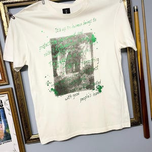 May include: Cream-coloured t-shirt with a crew neck, displaying a green and grey graphic print and text. The text includes phrases such as "It's up to human beings" and "protect". The t-shirt is suspended on a clothesline.