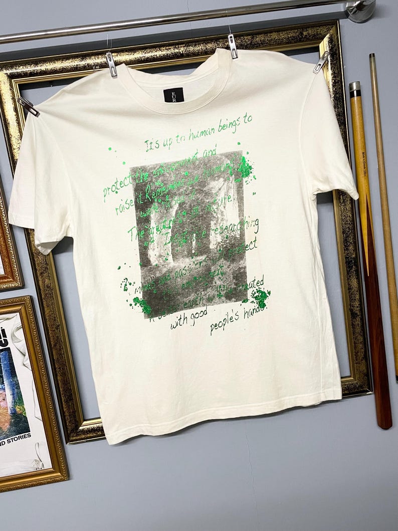 May include: Cream-coloured t-shirt with a crew neck, displaying a green and grey graphic print and text. The text includes phrases such as "It's up to human beings" and "protect". The t-shirt is suspended on a clothesline.
