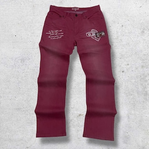 May include: Burgundy-colored jeans with a classic five-pocket design. Features white script on the left leg and a patch with the number "03" on the right. The jeans are laid flat against a textured background.