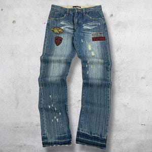 Pode incluir: Calças de ganga azuis com lavagem desgastada e aplicações decorativas. As calças de ganga apresentam um design clássico de cinco bolsos, fecho de botão e zíper e corte reto. As aplicações incluem um escudo, um oval e um retângulo.