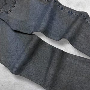 May include: Faded gray denim jeans with a classic five-pocket design. The jeans have a worn look, with visible stitching and a back pocket detail. The fabric appears soft and well-worn, suggesting comfort and a vintage aesthetic.