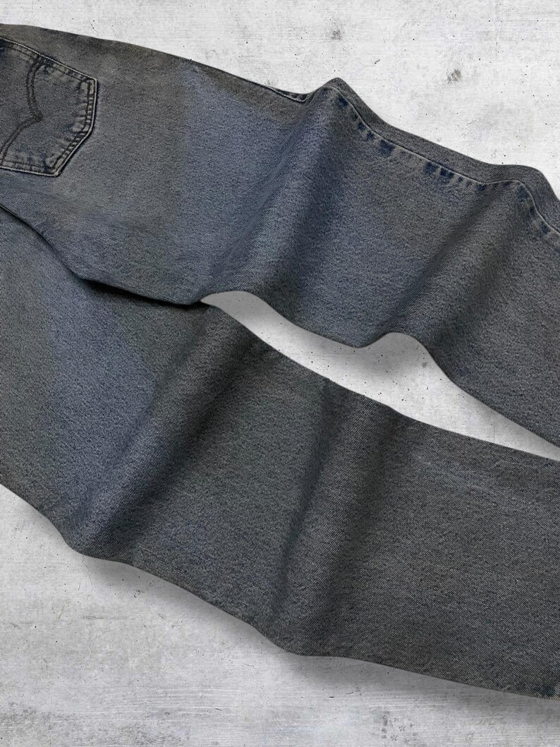 May include: Faded gray denim jeans with a classic five-pocket design. The jeans have a worn look, with visible stitching and a back pocket detail. The fabric appears soft and well-worn, suggesting comfort and a vintage aesthetic.