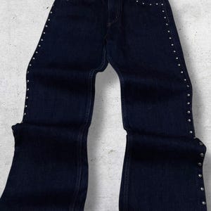 Puede incluir: Vaqueros de mezclilla azul oscuro con detalles de tachuelas plateadas a lo largo de las costuras laterales y los bolsillos. Los vaqueros tienen un dise&ntilde;o cl&aacute;sico de cinco bolsillos y una abertura acampanada en la pierna. Un b&aacute;sico vers&aacute;til del armario.