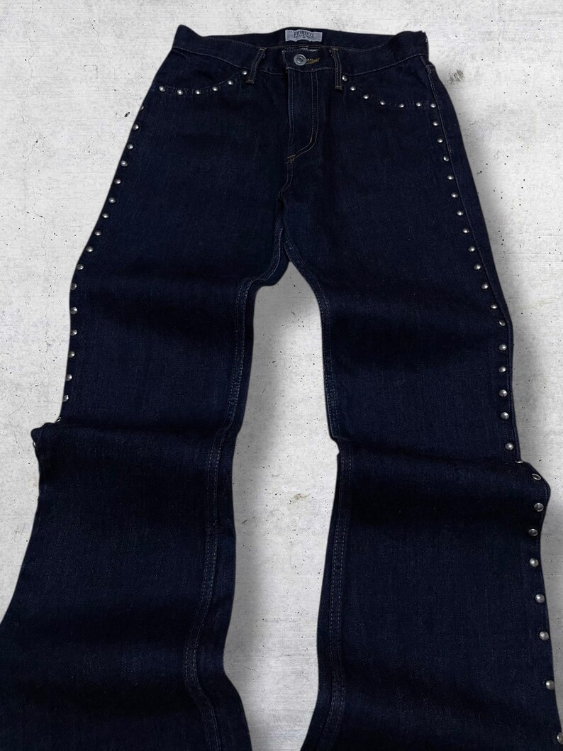 Puede incluir: Vaqueros de mezclilla azul oscuro con detalles de tachuelas plateadas a lo largo de las costuras laterales y los bolsillos. Los vaqueros tienen un dise&ntilde;o cl&aacute;sico de cinco bolsillos y una abertura acampanada en la pierna. Un b&aacute;sico vers&aacute;til del armario.