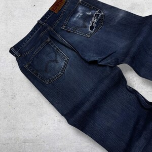 May include: Dark blue denim jeans with a distressed look, featuring a torn pocket and a leather patch with the brand name "EDWIN". The jeans have classic five-pocket styling and a faded appearance.