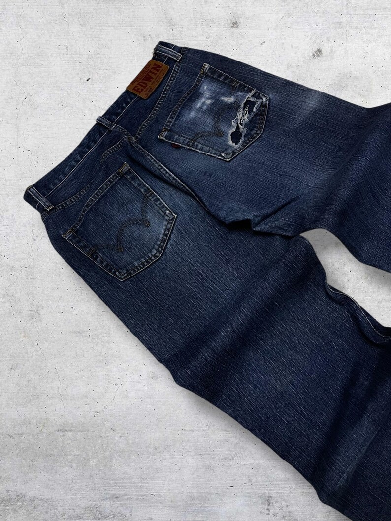 May include: Dark blue denim jeans with a distressed look, featuring a torn pocket and a leather patch with the brand name "EDWIN". The jeans have classic five-pocket styling and a faded appearance.