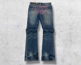 May include: Blue denim jeans with a flared leg. The back pockets feature pink embroidered text that reads "Christian Audigier". The jeans have a distressed look with faded areas and a worn hem.