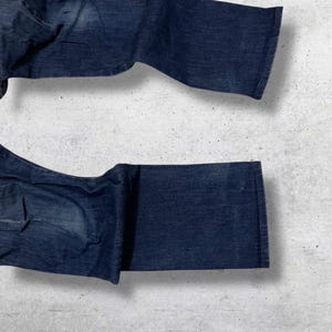 以下が含まれることがあります： 濃い青色のデニムジーンズ。脚の開口部と裾を見せるように折りたたまれています。ジーンズは、クラシックな5ポケットのデザインで、少し色あせた外観で、使い古された外観を示唆しています。生地は中厚手のデニムのようです。