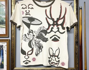 May include: Off-white t-shirt with a brown collar and sleeves, featuring a graphic design of Japanese Kabuki masks and floral elements in black and red. The shirt is hanging on a clothesline.