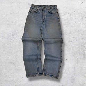 May include: Light blue denim jeans with a classic five-pocket design. The jeans have a faded wash and a straight leg cut. The waist is buttoned and the jeans are laid flat on a textured surface.