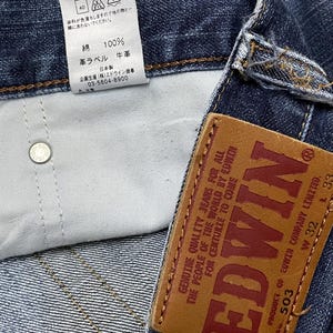 May include: Close-up of a pair of blue denim jeans with a leather brand tag. The tag reads "EDWIN" in large red letters. The jeans have a white tag with Japanese text and care instructions.