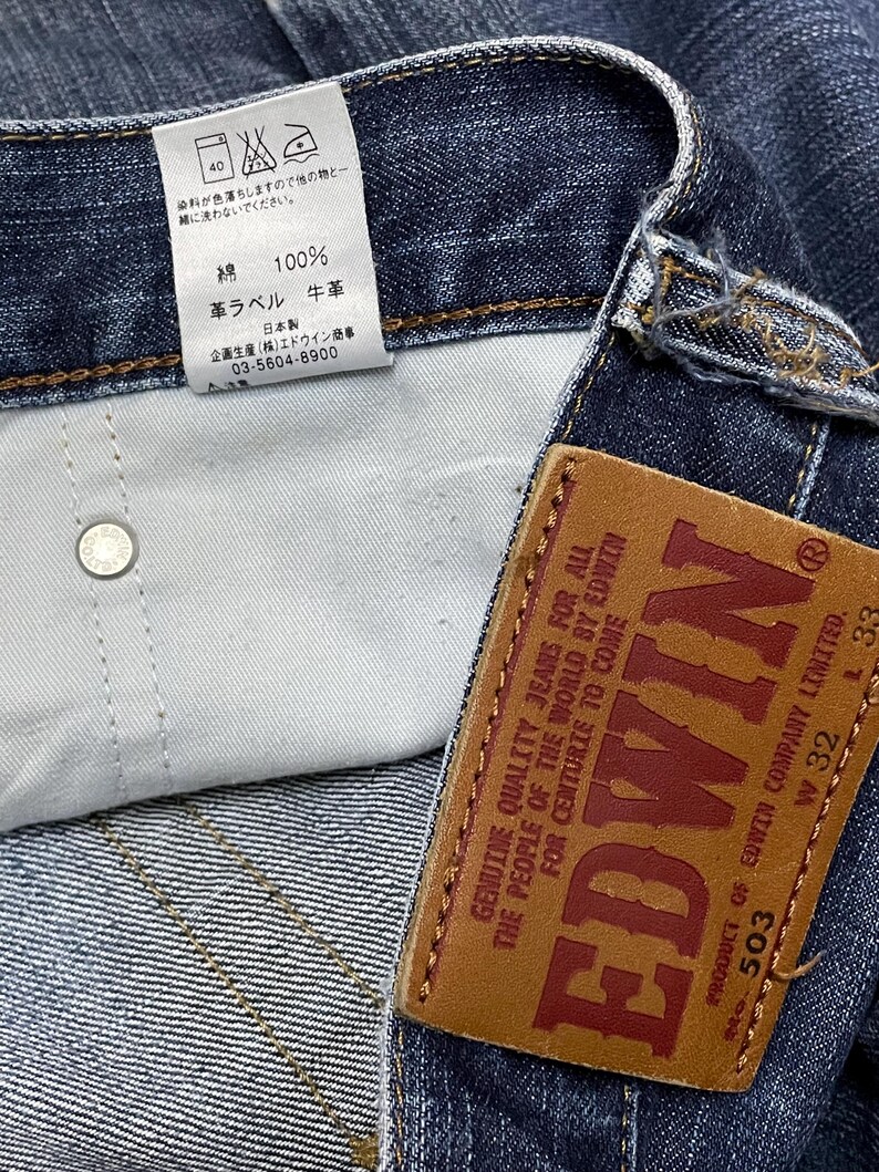 May include: Close-up of a pair of blue denim jeans with a leather brand tag. The tag reads "EDWIN" in large red letters. The jeans have a white tag with Japanese text and care instructions.