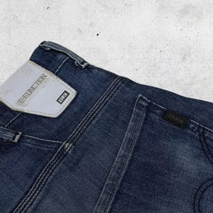 以下が含まれることがあります： 濃い青色のデニムジーンズのクローズアップ。ジーンズの背面、バックポケットとウエストバンドが見えます。「EDWIN」のブランド名がラベルに表示されています。ジーンズはクラシックで使い古された外観をしています。