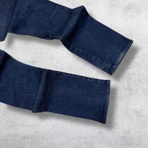 以下が含まれることがあります： 濃い青色のデニムジーンズ。ジーンズは折りたたまれ、明るい灰色の表面に置かれています。デニムはクラシックで、わずかに色あせた外観をしています。ジーンズは新品で未使用のようです。