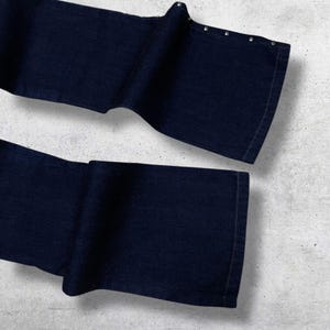 Puede incluir: Pantalones vaqueros azul marino oscuro con dise&ntilde;o de pernera ancha. Los pantalones tienen una serie de peque&ntilde;os remaches plateados a lo largo del borde superior de la cintura doblada. La tela parece ser un denim de peso medio.