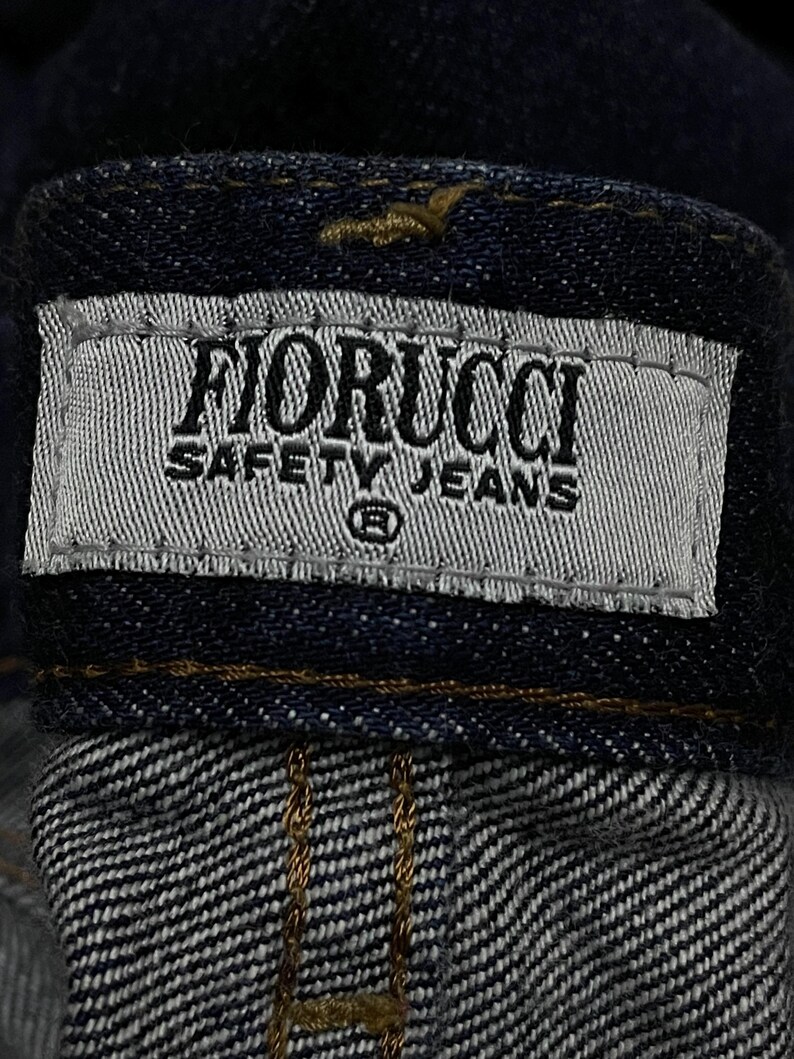 Puede incluir: Primer plano de una prenda de mezclilla azul oscuro, que muestra una etiqueta que dice "FIORUCCI SAFETY JEANS" en texto negro sobre un fondo gris claro. La etiqueta est&aacute; cosida a la mezclilla.