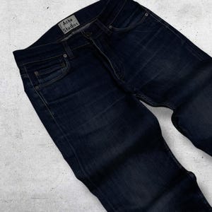 Op de afbeelding: Donkerblauwe denim jeans met een klassiek vijf-zakken ontwerp. De jeans hebben een vervaagde uitstraling en een kleine scheur bij de knie. Het merklabel "Acne Studios" is zichtbaar op de tailleband.