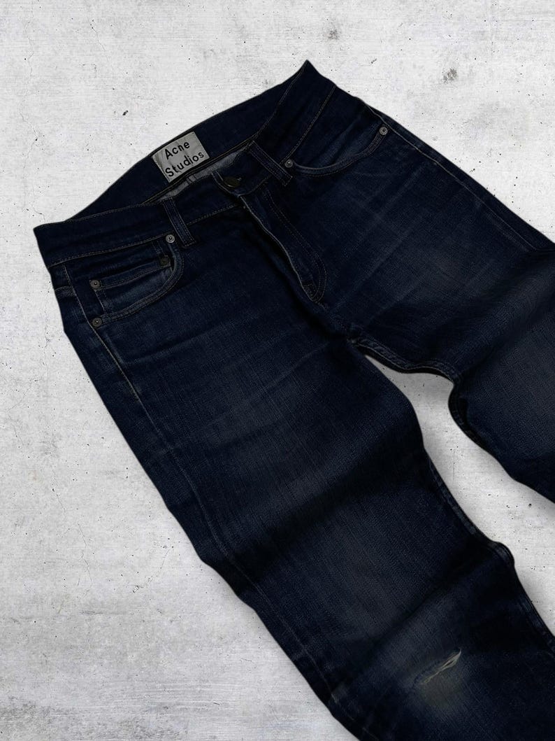 Op de afbeelding: Donkerblauwe denim jeans met een klassiek vijf-zakken ontwerp. De jeans hebben een vervaagde uitstraling en een kleine scheur bij de knie. Het merklabel "Acne Studios" is zichtbaar op de tailleband.