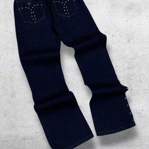 Puede incluir: Vaqueros de mezclilla azul oscuro con pernera acampanada. Los bolsillos traseros est&aacute;n adornados con tachuelas plateadas en un dise&ntilde;o estilizado. Tachuelas adicionales adornan las costuras exteriores cerca de los tobillos. Los vaqueros est&aacute;n sobre una superficie gris claro.