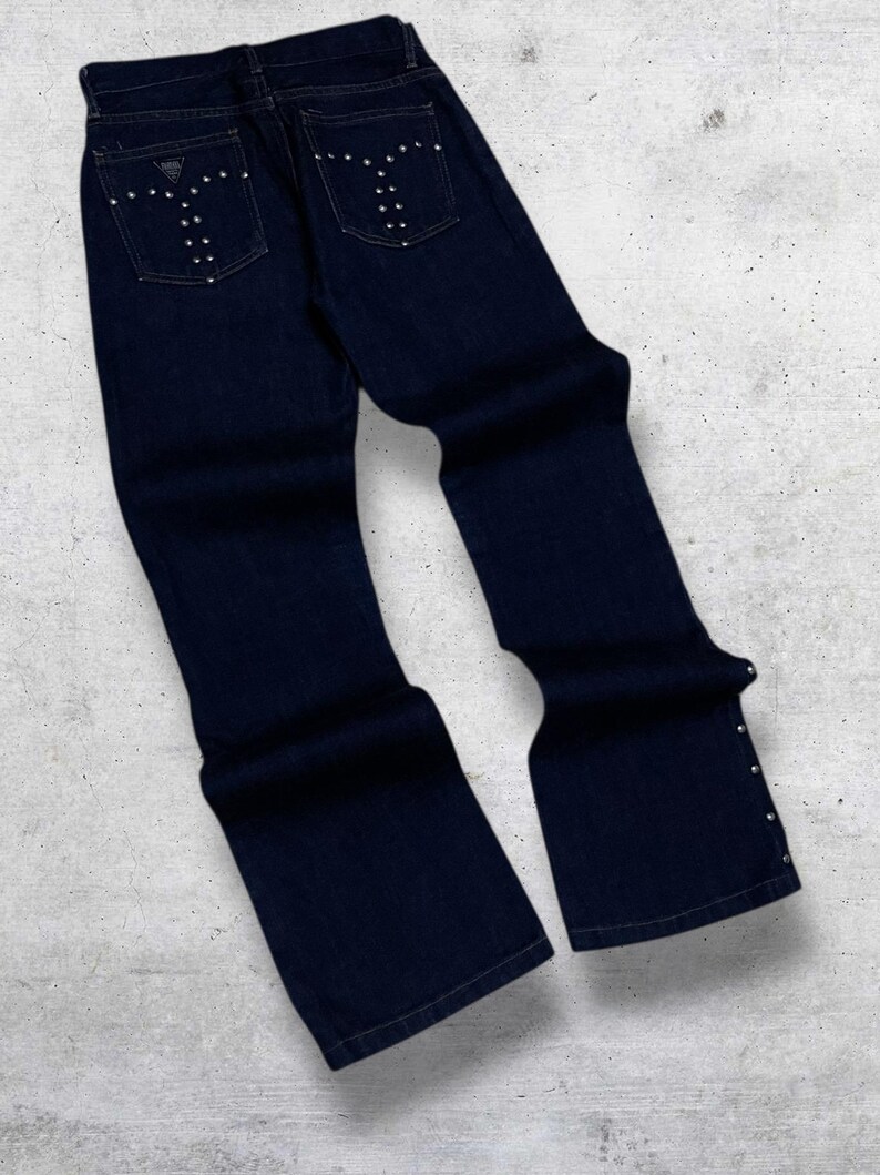 Puede incluir: Vaqueros de mezclilla azul oscuro con pernera acampanada. Los bolsillos traseros est&aacute;n adornados con tachuelas plateadas en un dise&ntilde;o estilizado. Tachuelas adicionales adornan las costuras exteriores cerca de los tobillos. Los vaqueros est&aacute;n sobre una superficie gris claro.