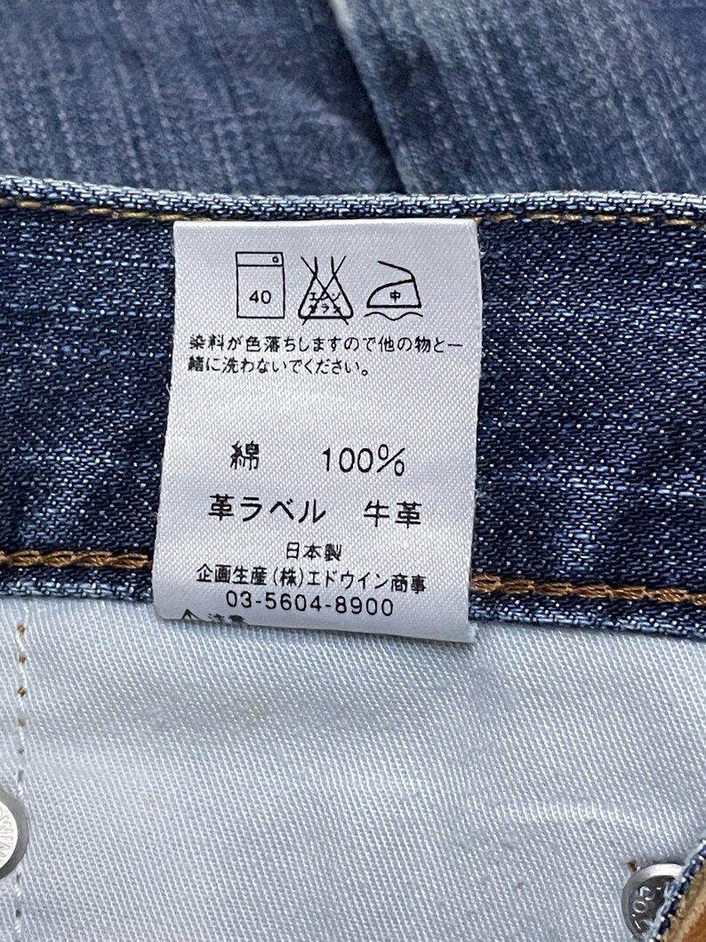 May include: Close-up of a denim jeans label with Japanese text and care symbols. The label indicates 100% cotton, leather, and washing instructions. The label also includes a phone number.