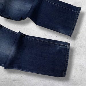 May include: Dark wash denim jeans, folded and laid out on a light gray surface. The jeans have a classic five-pocket design and a straight leg cut. The denim has subtle fading and wear marks.