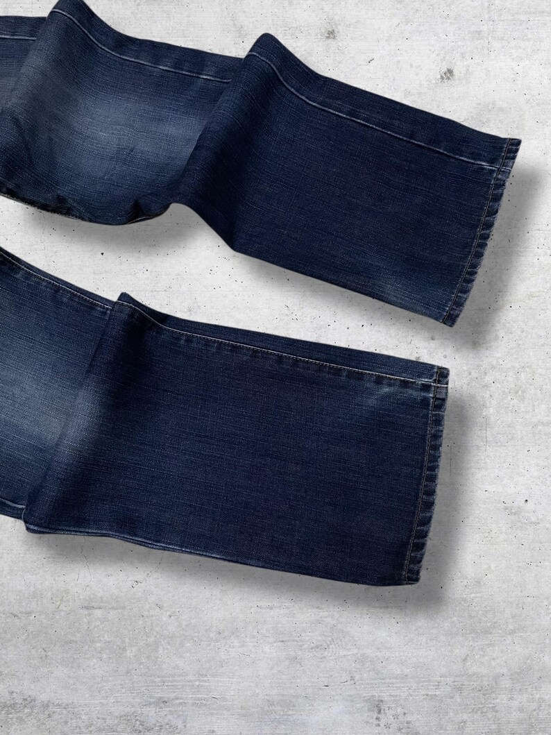 May include: Dark wash denim jeans, folded and laid out on a light gray surface. The jeans have a classic five-pocket design and a straight leg cut. The denim has subtle fading and wear marks.