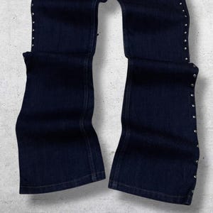 Puede incluir: Pantalones vaqueros azul oscuro con dise&ntilde;o de pernera acampanada. Los lados de los pantalones est&aacute;n adornados con peque&ntilde;os tachuelas plateadas. Los pantalones est&aacute;n colocados sobre una superficie gris claro.