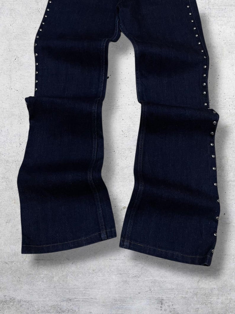 Puede incluir: Pantalones vaqueros azul oscuro con dise&ntilde;o de pernera acampanada. Los lados de los pantalones est&aacute;n adornados con peque&ntilde;os tachuelas plateadas. Los pantalones est&aacute;n colocados sobre una superficie gris claro.