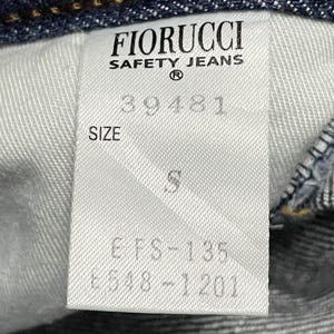 Puede incluir: Primer plano de una etiqueta blanca de unos vaqueros Fiorucci Safety. La etiqueta muestra el nombre de la marca, la talla y los c&oacute;digos de producto. Los vaqueros son de mezclilla azul oscuro.