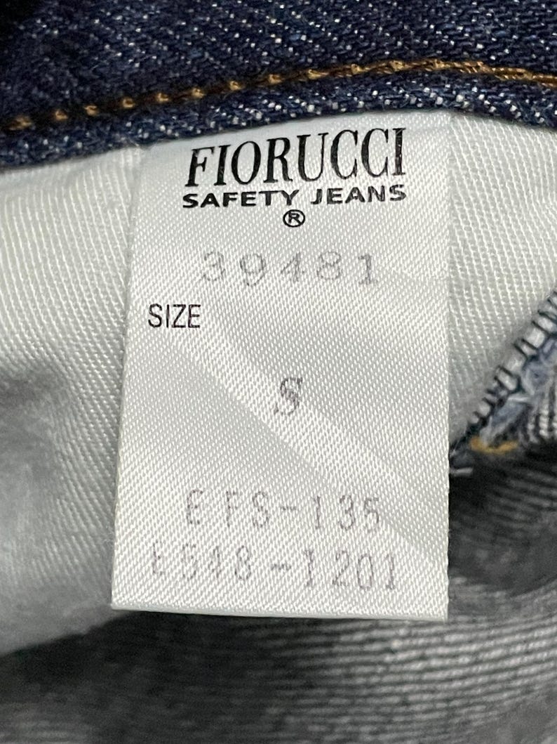 Puede incluir: Primer plano de una etiqueta blanca de unos vaqueros Fiorucci Safety. La etiqueta muestra el nombre de la marca, la talla y los c&oacute;digos de producto. Los vaqueros son de mezclilla azul oscuro.