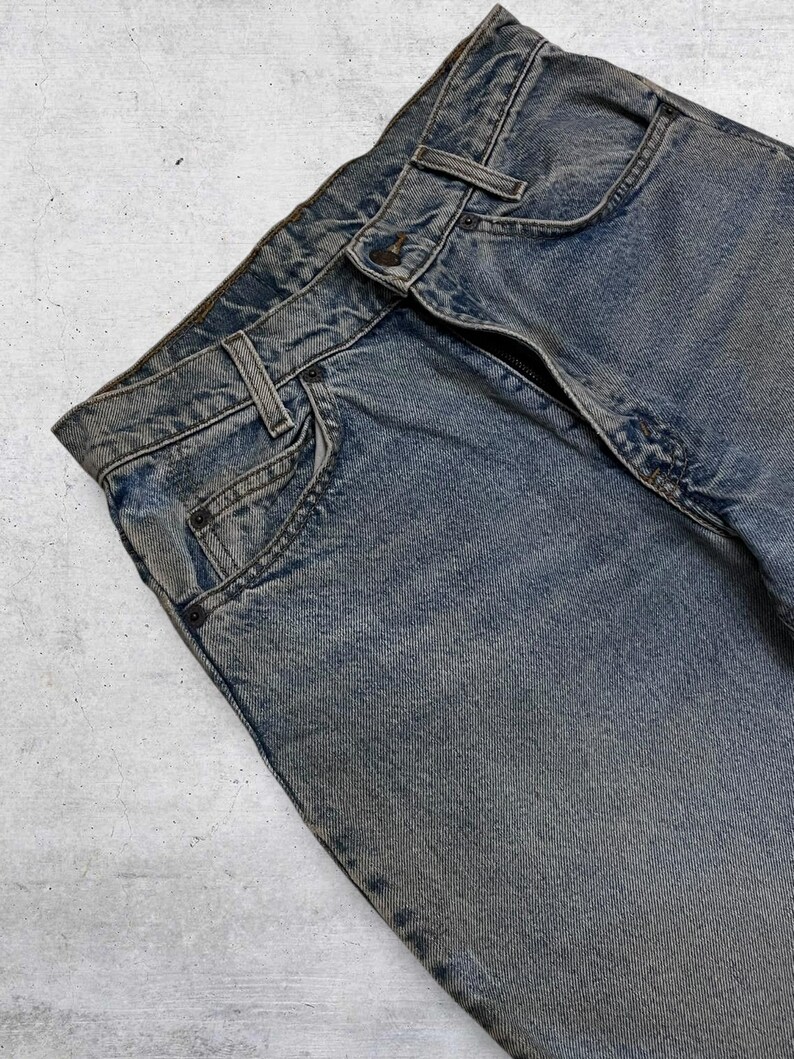 May include: Close-up of faded blue denim jeans. The jeans have a button closure, belt loops, and classic five-pocket styling. The denim has a worn, textured appearance, with subtle variations in color.