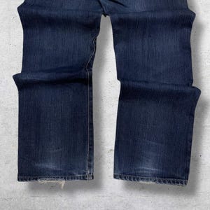 May include: Dark blue denim jeans with a classic five-pocket design. The jeans have a straight leg cut and show slight wear at the hem. The fabric has a textured appearance.