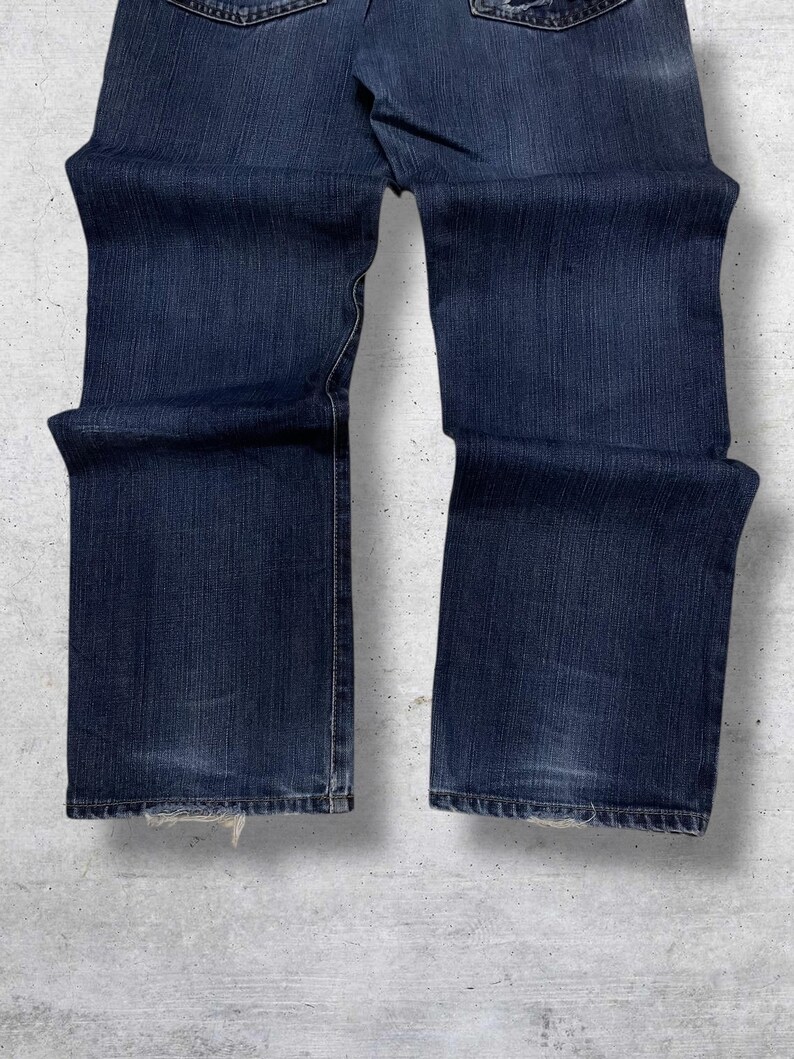May include: Dark blue denim jeans with a classic five-pocket design. The jeans have a straight leg cut and show slight wear at the hem. The fabric has a textured appearance.